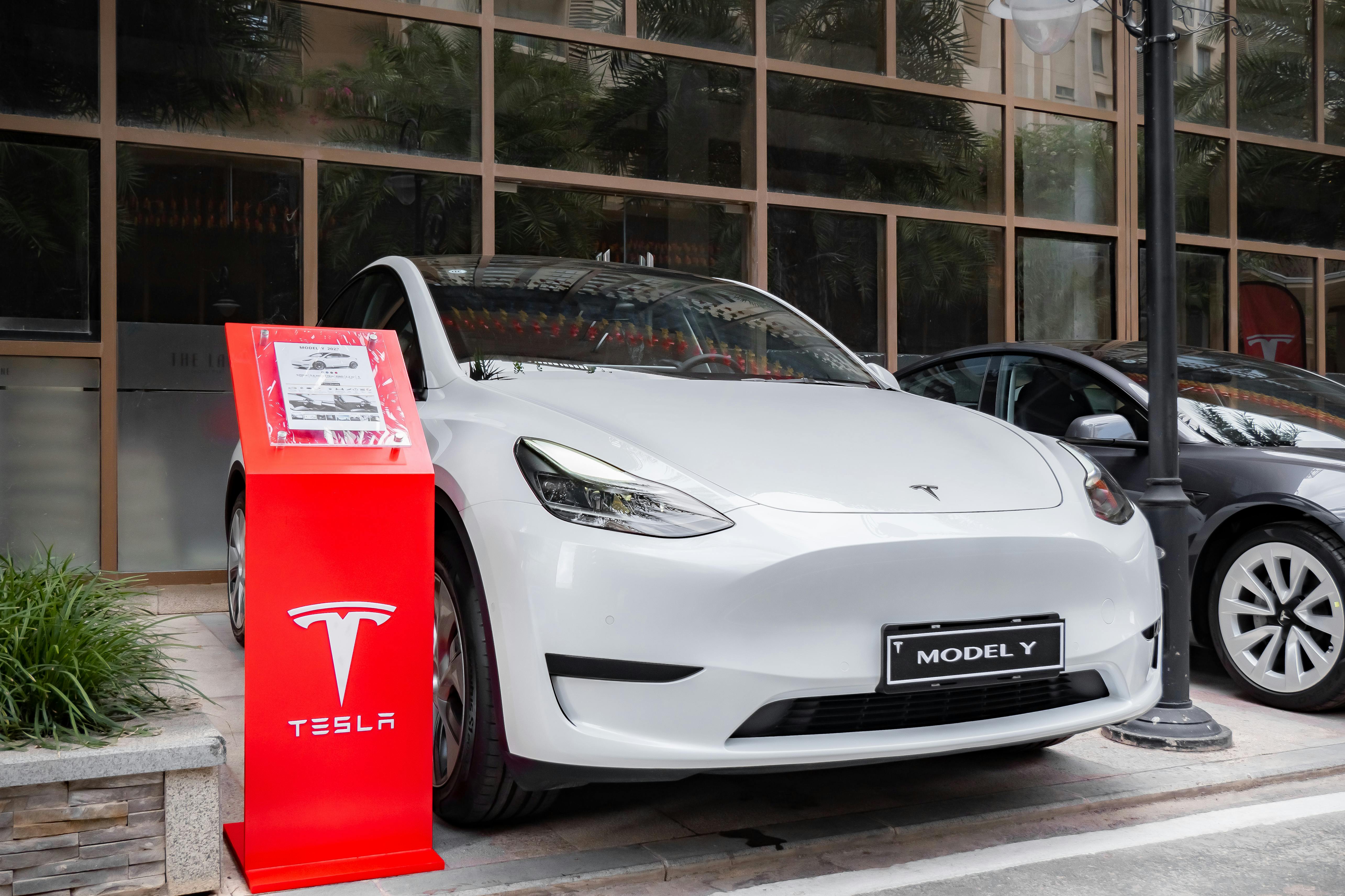 White Tesla Model Y with a red Tesla lectern next to it, windows behind and a partial black ground to the right